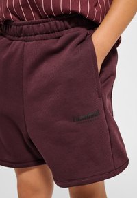 Burgundy shorts med elastisk midjeband, med sidofickor och en svart logodetalj på vänster ben. Slät, blandning av bomull.