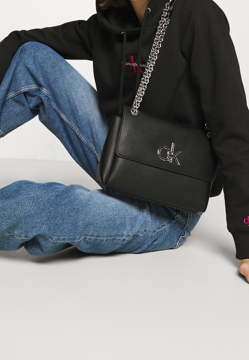 Sort læderhåndtaske med en sølvfarvet kædetræksrem, der har et metallogo-detalje på forsiden og et rent, struktureret design.