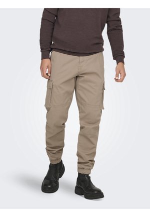 Mand iført beige cargobukser med elastiske manchetter, sorte støvler og en mørkebrun ribstrikket sweater, stående mod en lys baggrund.