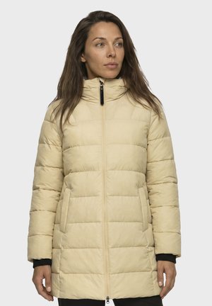 Frau mit langen braunen Haaren, die eine beige gesteppte Daunenjacke mit Reißverschluss trägt, schaut leicht nach rechts vor einem einfachen hellen Hintergrund.