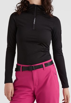 Žena nosící přiléhavý černý top s dlouhým rukávem a předním zipem a růžové kalhoty s černým páskem, pózující s uvolněnýma rukama.