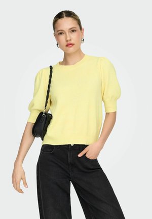 Donna che indossa un maglione giallo con maniche a sbuffo, jeans neri, orecchini a cerchio argentati e porta una borsa a tracolla nera.