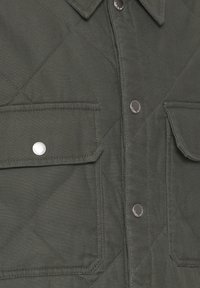 Prošívaná tmavě zelená košile s kabelkou vyrobená z texturované látky, s dvěma prsními kapsami, stříbrnými patentkami a formovaným límečkem.