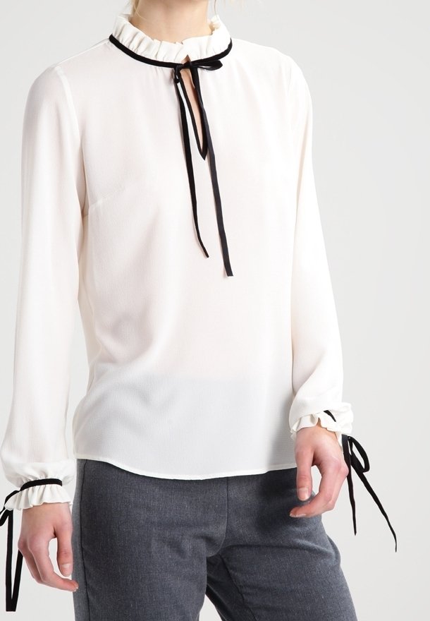 Femme portant un chemisier blanc à manches longues avec un col à volants et des rubans noirs au niveau du cou et des poignets, associé à un pantalon gris.