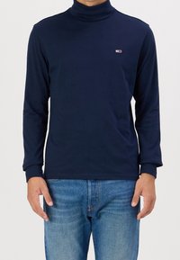 Navyblaues Rollkragenhemd aus Baumwolle mit langen Ärmeln, das ein kleines Logo auf der Brust und eine taillierte Passform aufweist. Getragen mit blauen Jeans.