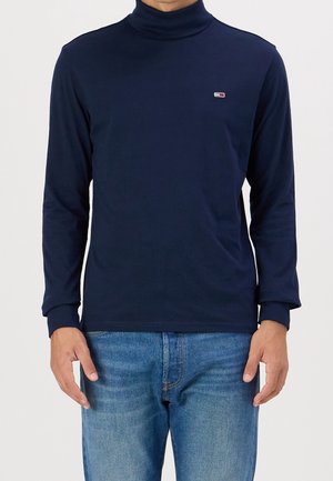 Marinblå polotröja i bomull med långa ärmar, med en liten logotyp på bröstet och en figurnära design. Bärs med blå jeans.