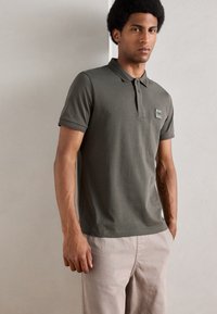 Tricou polo de culoare verde măslin, cu un guler texturat și o mică emblema, asortat cu pantaloni de culoare deschisă; material din bumbac neted.