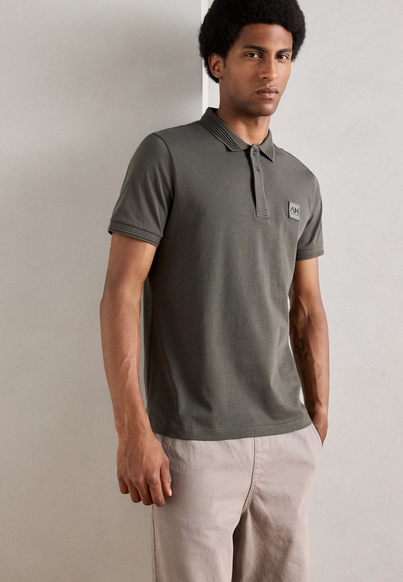 Tricou polo de culoare verde măslin, cu un guler texturat și o mică emblema, asortat cu pantaloni de culoare deschisă; material din bumbac neted.