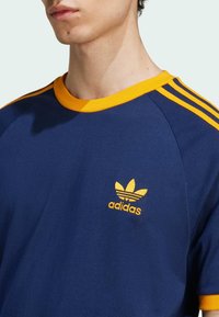 Navy blue t-shirt with yellow accents, featuring a crew neckline and three yellow stripes on the shoulders. Embroidered Adidas logo on chest.