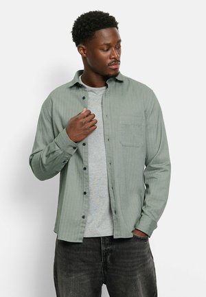 Light green, striped long-sleeve shirt with a button-up front, single chest pocket, and dark buttons, worn over a gray t-shirt and dark jeans.