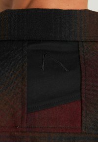 Primo piano di un colletto di giacca in lana multicolore con una toppa di tessuto nero e un logo ricamato sottile sul retro del collo.