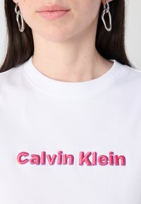 Balts kokvilnas T-kreklu ar izšūtu tekstu "Calvin Klein" rozā gradientā. Apkakle ir ribota, ar gludu auduma faktūru.