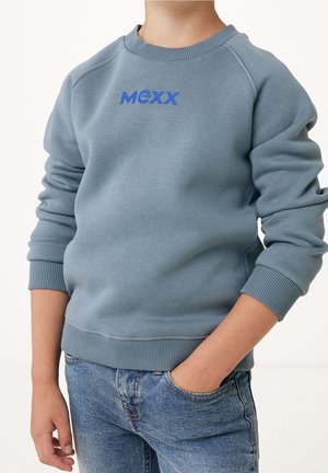Kind trägt ein hellblaues MEXX-Sweatshirt mit langen Ärmeln und blaue Jeans aus Denim, eine Hand in der Tasche vor einfarbigem Hintergrund.