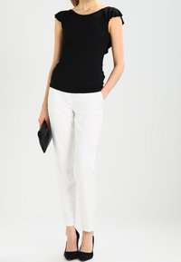 Žena v černém topu s volány a bílých kalhotách, držící černou kabelku, stojící s jednou rukou v kapse a na černých lodičkách.