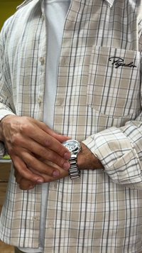 Camicia beige a quadri con bottoni, linee grigie e una tasca. Orologio argento con cinturino in metallo al polso. Maglietta bianca visibile sotto.