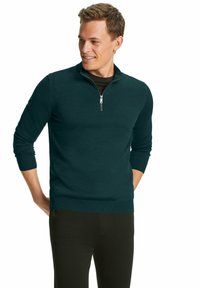 Maglione verde acqua con collo a zip in un morbido tessuto testurizzato, maniche lunghe e polsini a costine. Abbinato a pantaloni neri, presenta un design aderente.