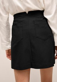 Minifalda negra con cintura ajustada, dos bolsillos traseros y una textura suave. El dobladillo es ligeramente curvado. Combinada con una blusa blanca.