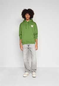 Green hoodie with a front pocket and drawstring hood, featuring a white logo. Paired with light gray jeans and white sneakers.