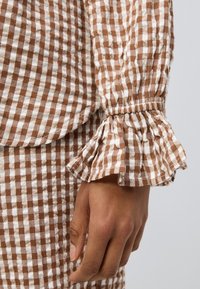 Ύφασμα με καφέ και λευκό καρό και βολάν στους μανσέτες. Το υλικό φαίνεται ελαφρύ και με υφή, παρουσιάζοντας ένα casual μοτίβο και σχέδιο.