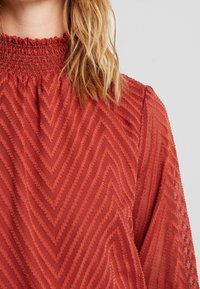 Rote strukturierte Bluse mit hohem Rüschenausschnitt und Chevron-Muster. Der Stoff ist leicht und semi-transparent.