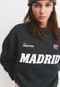 Čierna mikina s veľkým bielym textom "MADRID", malým ružovým textom "nový KREATÍVNY TÍM" a "1991", s rebrovaným golierom a manžetami.
