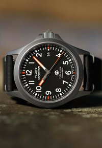 Montre Timex Expedition avec un cadran noir, un boîtier en titane, des indices blancs et orange, et un bracelet en cuir noir ; dotée de aiguilles luminescentes.