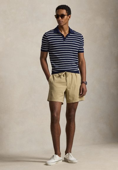 Άνδρας που φοράει μπλε ναυτικό ριγέ πόλο με κοντά μανίκια, μπεζ σορτς με κορδόνι, λευκά αθλητικά παπούτσια, γυαλιά ηλίου και ρολόι χειρός, στέκεται με τα χέρια στις τσέπες.