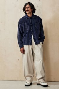 Cămașă overshirt de corduroy albastru închis cu închidere cu nasturi și două buzunare la piept, asortată cu pantaloni largi bej deschis și pantofi albi cu tălpi negre.