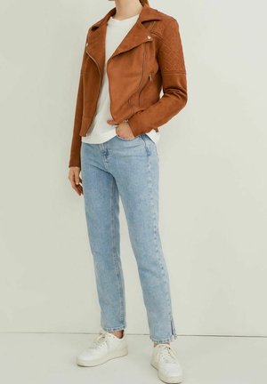 Person iført en brun quiltet læderjakke, hvid skjorte, lyseblå jeans med ankelslids, og hvide sneakers, stående mod en ensfarvet baggrund.