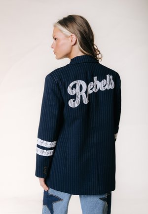 Donna con capelli castano chiaro che indossa un blazer blu navy a righe con strisce bianche sulle maniche e "Rebels" ricamato sulla schiena.