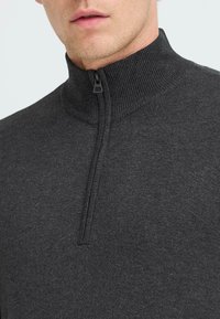 Suéter gris oscuro con un alto cuello y cierre de media cremallera. Fabricado en un tejido suave y texturizado; costuras visibles cerca de la cremallera.