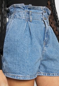 Hoch geschnittene Jeansshorts in Blau mit Falten, zwei Knöpfen und einem gesteppten Saum. Der Stoff hat eine strukturierte Optik mit einem leichten Glanz.