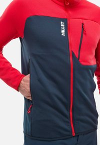 Fleece-zipjacke, rotes Oberteil und marineblaues Unterteil, verfügt über eine Brusttasche, Seitentaschen und eine strukturierte Oberfläche. Logo auf der Vorderseite angebracht.