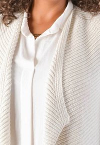 Knit cardigan in off-white with ribbed texture, open front, layered over a white collared shirt. Simple, elegant design without embellishments.