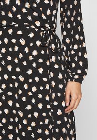 Robe portefeuille noire à manches longues avec des taches abstraites blanches et beiges éparses, nouée à la taille avec une ceinture fine, main détendue sur le côté.