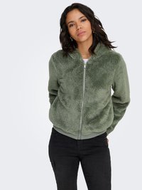 Veste zippée verte duveteuse avec un col montant, des poignets et un ourlet côtelés. Associée à un pantalon foncé, le tissu est doux au toucher.