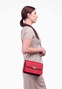 Červená kožená křížová taška se zlatými kovovými prvky, obdélníkového tvaru, s klopovým zavíráním. Design obsahuje decentně embosované logo na přední straně.