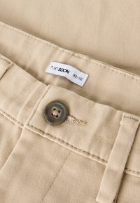 Pantaloni di cotone beige con una texture liscia, dotati di un bottone grigio e una cintura cucita. L'etichetta riporta "TWO SOON 32/32."