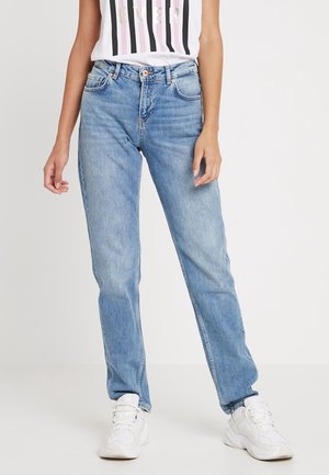 Femme portant un jean droit bleu clair, des baskets blanches épaisses et un t-shirt blanc sans manches à rayures verticales.