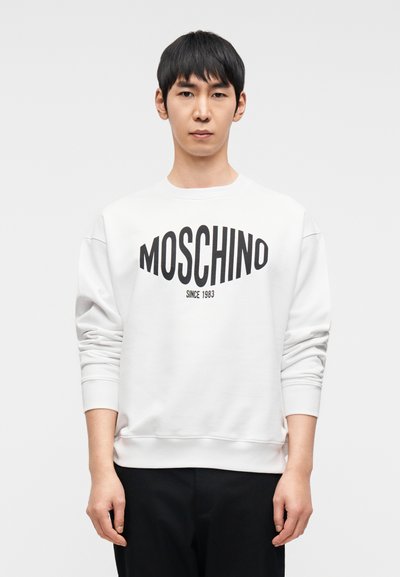 Λευκή φούτερ με μαύρο λογότυπο "MOSCHINO", στρογγυλή λαιμόκοψη, μακριά μανίκια και ρεβέρ στα μανίκια. Φορεμένη πάνω από μαύρο παντελόνι. Απλό φόντο.