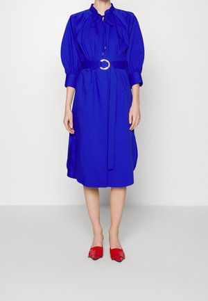 Mujer con un vestido azul cobalto hasta la rodilla con mangas abullonadas y un cinturón a juego, combinado con zapatos planos rojos de punta afilada.