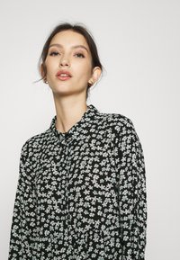 Vestido camisola preto de mangas longas com padrões florais brancos e azul claro, colarinho arredondado e fecho de botão na parte da frente.