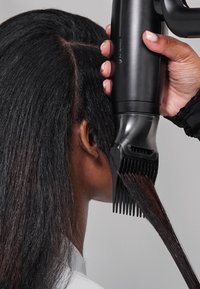 Cheveux d'une personne lissés à l'aide d'une brosse chauffante noire tenue par une main aux ongles vernis en rose clair.