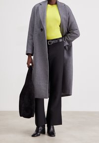 Grey wool coat, bright yellow knit top, black trousers, and black heeled boots. Holding a black suede bag. Modern, long silhouette.