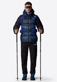 Niebieska i czarna izolowana kurtka z kapturem, z zamkiem z przodu, paski z przodu i kieszeniami bocznymi; czarne spodnie; niebieskie buty trekkingowe; kijki trekkingowe.