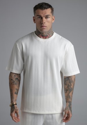 Man with neck and arm tattoos wearing a textured white short-sleeve shirt and matching pants against a plain gray background.