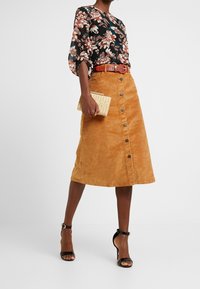 Czarna bluzka w kwiaty z bufiastymi rękawami, pomarańczowa zamszowa spódnica midi z przodem na guziki oraz pleciona beżowa torebka kopertówka. Czarne sandały na wysokim obcasie.