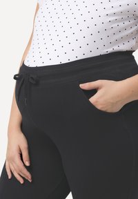 Schwarze flexible Hose mit einem elastischen Bund und Kordelzug sowie seitlichen Taschen, kombiniert mit einem weißen Oberteil mit Punkten.