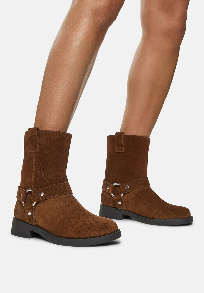 Botas de ante marrón hasta el tobillo con punta redonda, suela negra plana y un acento de hardware plateado en una correa alrededor del tobillo.