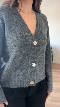 Grå strikket cardigan med V-hals, fem knapper (tre dekorative, to funksjonelle), ribbestrikket kant og lange ermer. Myk tekstur synlig.